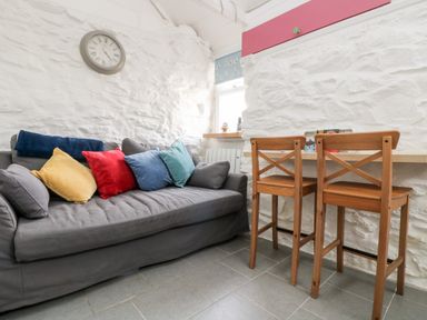 A small living area with a grey sofa with colorful cushions a wall clock wooden chairs and a narrow wooden table at The Old Stable in Porthallow