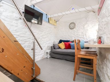 A room with a gray sofa with colorful cushions a wall clock a wooden chair a mounted TV and wooden stairs at The Old Stable Porthallow