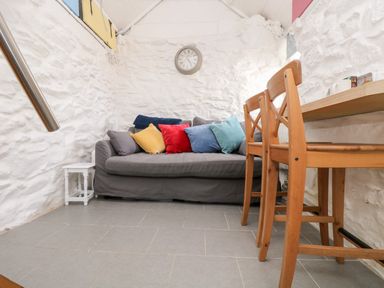 A room with a gray sofa with colorful cushions a wooden table with chairs and white stone walls at The Old Stable in Porthallow