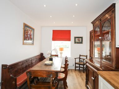 A dining room with wooden chairs a wooden table a bench with red cushions a window with a red blind and a wooden cabinet at Bryn Afon Farm in Llanrug