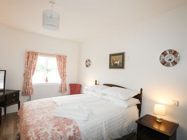 A bedroom with a bed with white linens and folded towels a nightstand with a lamp a window with patterned curtains a chair and decorative plates on the wall at Bryn Afon Farm Llanrug