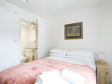 A bedroom with a bed covered with a pink blanket and white pillows a wooden nightstand with a lamp a door open to a bathroom with a sink and toilet a framed picture on the wall at Bryn Afon Farm in Llanrug