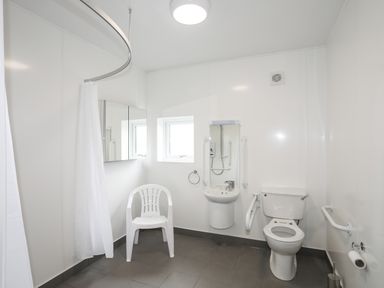 A bathroom with a toilet, sink, white plastic chair, shower curtain, and safety rails at Bryn Afon Farm in Llanrug