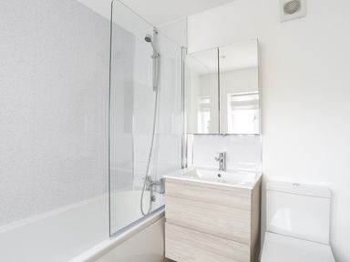 A bathroom with a bathtub shower, a sink with a wooden cabinet, a mirrored medicine cabinet, and a toilet at Bryn Afon Farm in Llanrug