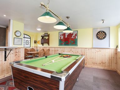 A room with a pool table billiard cues and balls a dartboard a table with four chairs and framed pictures on yellow walls at Bryn Afon Farm in Llanrug