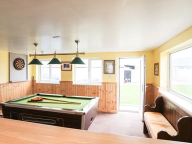 A game room with a pool table and dartboard next to windows and an open door with a bench along the wall at Bryn Afon Farm in Llanrug