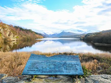 A lake with surrounding trees and snow-capped mountains in the background with an information plaque in the foreground at Bryn Afon Farm in Llanrug