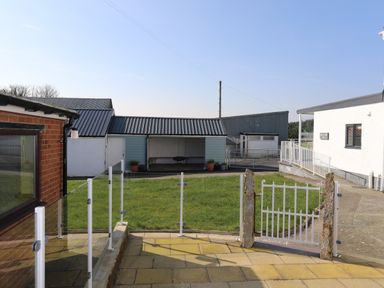 An outdoor area with a small grassy yard, a white metal gate, glass fences, and various buildings at Bryn Afon Farm in Llanrug