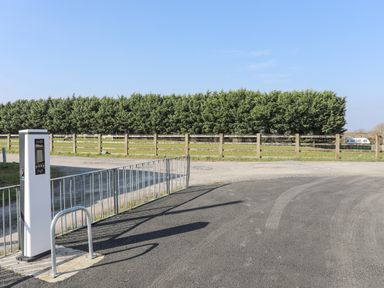 An electric vehicle charging station near a fenced grassy field with trees in the background at Bryn Afon Farm in Llanrug