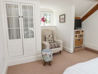 A corner of a bedroom with an armchair, a small floral stool, a cabinet with a TV, and a window at Garden Cottage in Garndolbenmaen near Tremadog