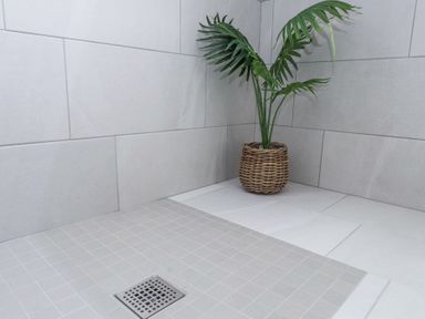 A corner of a tiled bathroom with a floor drain and a potted plant in a wicker basket