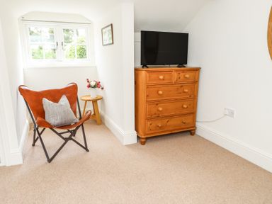 A small room with a leather chair a cushion a wooden side table with flowers a window and a wooden chest of drawers with a TV on top at Garden Cottage in Garndolbenmaen near Tremadog