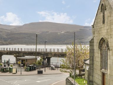 A street with a stone building on the right and a bridge over a body of water with hills in the background at Sapphire Suite in Barmouth