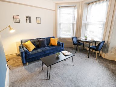 A living room with a blue sofa yellow and blue cushions a black coffee table and a small dining table with two chairs at Sapphire Suite in Barmouth