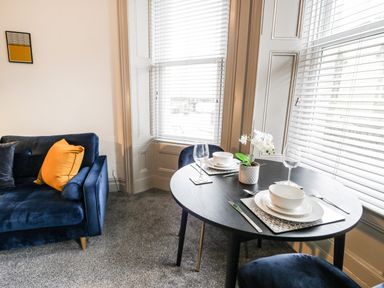 A dining table set for two with white dishes and wine glasses next to a blue sofa with pillows at Sapphire Suite in Barmouth
