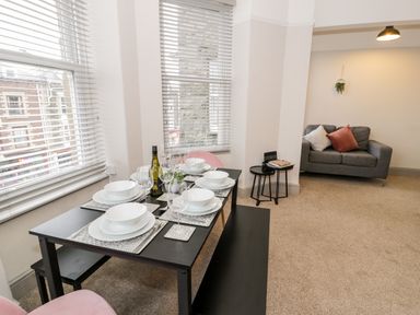 A dining table set with plates glasses and a bottle next to windows with blinds and a gray sofa with cushions in the background at Rose Suite in Barmouth