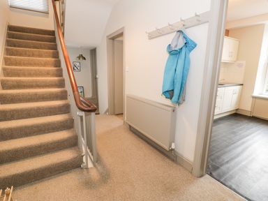 A hallway with carpeted stairs a blue jacket hanging on a wall hook and a view into a kitchen with black flooring at Rose Suite in Barmouth