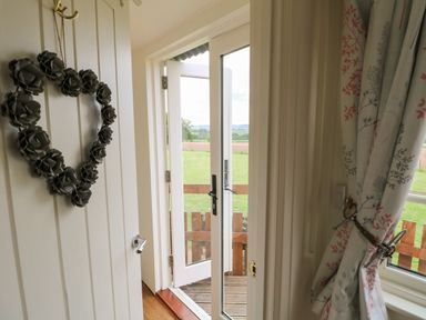 An open door leading to a wooden deck with a grassy field beyond a window with floral curtains at Shepherds Hut in Colton near Rugeley Staffordshire