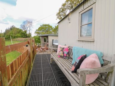 An outdoor seating area with a wooden bench and cushions next to a small fenced yard at Shepherds Hut in Colton near Rugeley Staffordshire