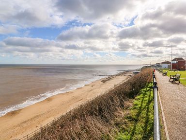 A sandy beach next to a grassy promenade with benches and houses at The Nook in Mundesley