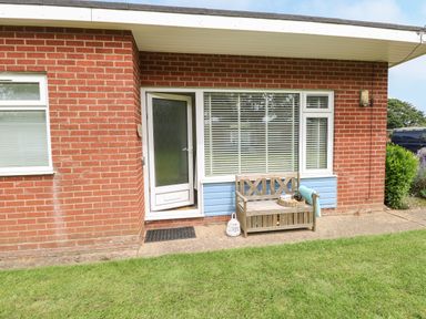 A brick exterior with a glass door next to a window and a wooden bench on a concrete path with grass in front at The Nook in Mundesley