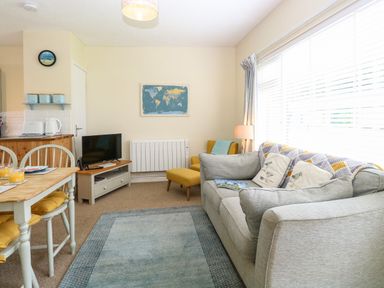 A living room with a gray sofa and cushions a small TV on a stand a yellow armchair a wooden dining table with chairs and cushions a world map wall art and large windows with blinds at The Nook in Mundesley