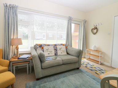 A living room with a gray sofa, a yellow armchair, side tables, a lamp, curtains, and a rug at The Nook in Mundesley