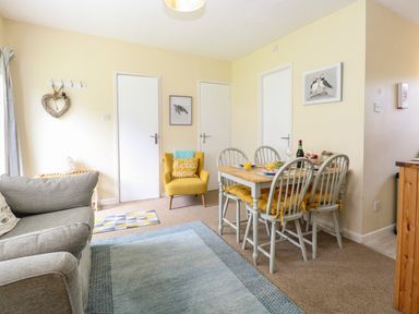 A dining area with a wooden table and four chairs with yellow cushions next to a yellow armchair and a gray sofa in a living room at The Nook in Mundesley