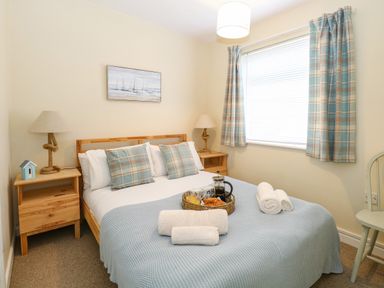 A bedroom with a bed covered with a blue blanket and pillows two nightstands with lamps a chair and a window with plaid curtains at The Nook in Mundesley