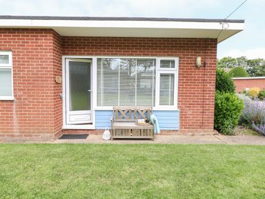 Front of a brick house with a white door and window a wooden bench and green lawn at The Nook in Mundesley