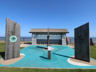 A coastal viewing shelter with wooden panels and a central wooden sculpture on a blue painted ground at The Nook in Mundesley