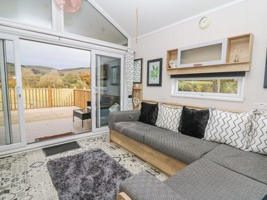 A living area with a large gray sectional sofa with black and white pillows a small rectangular rug a patterned floor a sliding glass door leading to a wooden deck and countryside view at Hares Rest in Nantmel near Rhayader