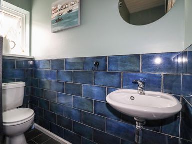 A bathroom with a toilet sink round mirror blue tiled walls and a window with a pitcher and reed diffuser on the sill at Ocean View in Downderry