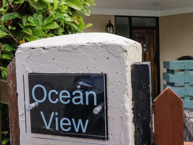 A sign reading Ocean View on a white concrete post near a wooden door and green plants at Ocean View in Downderry