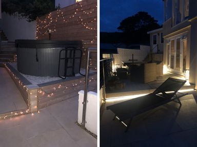 An outdoor patio area at night with a hot tub wrapped in string lights and a seating area with wicker furniture and a lounge chair at Ocean View in Downderry