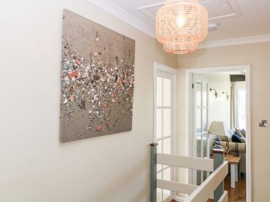 A hallway with a textured artwork on the wall a wicker pendant light and a view into a living room showing a striped sofa and table lamp at Ocean View in Downderry