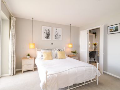 A bedroom with a white bed and yellow cushions two bedside tables with plants two hanging lamps and an open door showing a chair and sunflower vase at Ocean View in Downderry