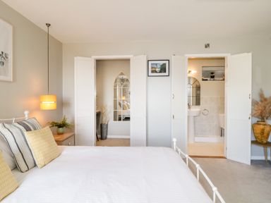 A bedroom with a bed white bedding yellow and striped pillows side table with plant hanging lamp and open doors to bathroom and another room at Ocean View in Downderry
