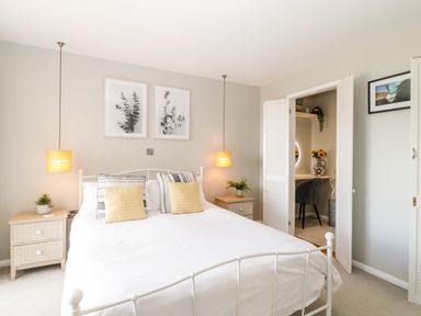 A bedroom with a white bed, bedside tables, hanging lamps, framed pictures, and an open closet with a desk at Ocean View in Downderry