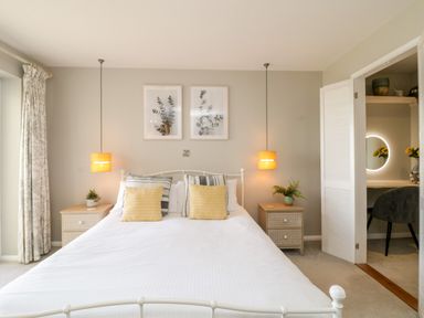 A bedroom with a bed with pillows bedside tables with lamps and plants and a small room with a chair and round mirror Ocean View Downderry