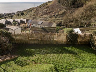 A grassy backyard with a wooden lattice fence overlooking houses and a hillside near the sea at Ocean View in Downderry