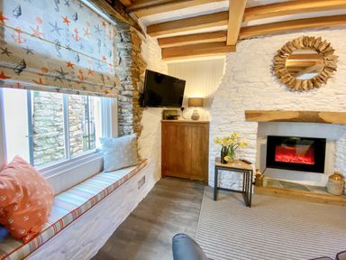 A sitting area with a window seat cushions a tv on a wooden cabinet a table with flowers and a fireplace in Pilchard Cottage in Brixham