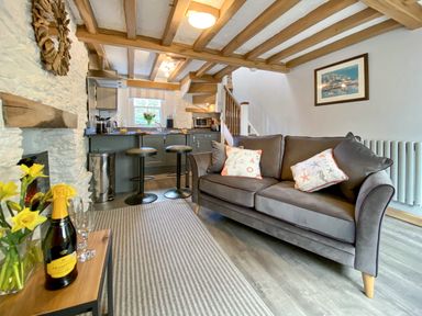 A living room with a gray sofa and a kitchen area with bar stools and wooden beams at Pilchard Cottage in Brixham