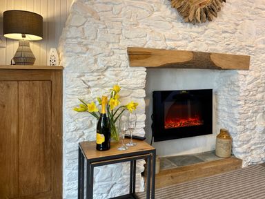 A fireplace with a wooden mantel and electric fire with a small table holding a bottle of prosecco and two glasses next to a cabinet with a lamp and yellow flowers at Pilchard Cottage in Brixham