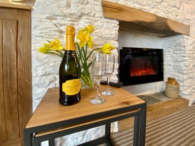 A wooden table with a bottle of prosecco and two glasses next to a vase of yellow flowers by a fireplace at Pilchard Cottage in Brixham