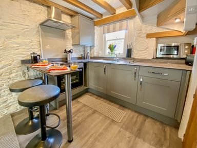 A kitchen with grey cabinets a breakfast bar with two stools and a window above the sink at Pilchard Cottage in Brixham
