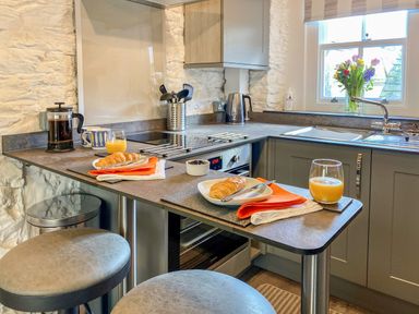A kitchen counter with two plates of croissants orange napkins juice glasses a coffee press kettle and a window with flowers at Pilchard Cottage in Brixham