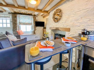A kitchen area with a breakfast bar set for two with orange juice and bread rolls a sofa and window seat with cushions and a TV mounted on a wall at Pilchard Cottage in Brixham