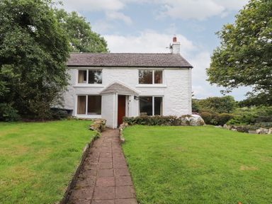 A white two story house with a tiled roof and four windows with a paved path leading to the entrance surrounded by grass and trees at Penllain in Newport Pembrokeshire