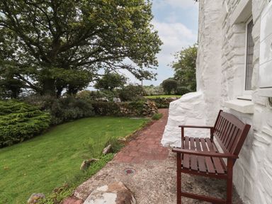 A wooden bench beside a white stone wall on a paved pathway with a grassy garden and trees at Penllain in Newport Pembrokeshire